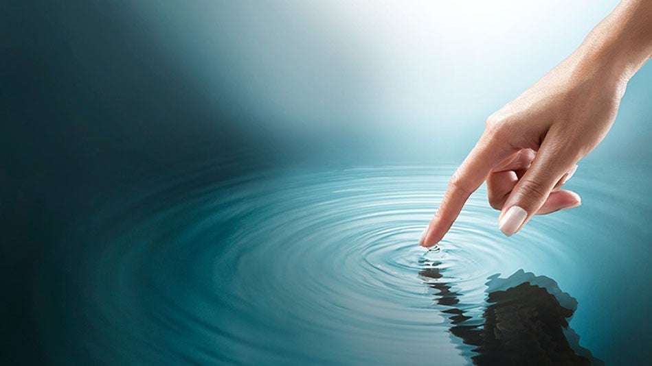 A hand with the index finger extended touching the surface of a body of water and causing a ripple.