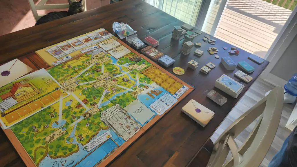 The Stardew Valley board game and all of its game pieces on a table with a cat looking on from nearby.