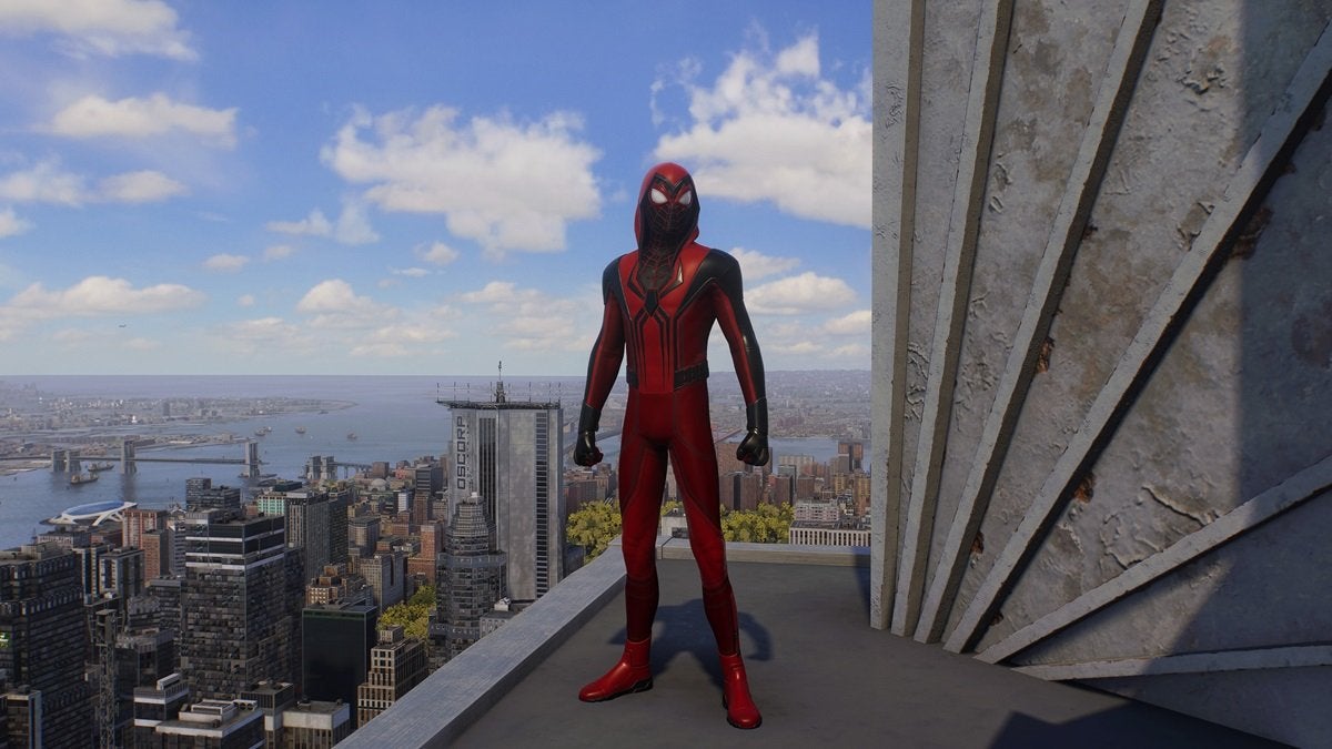 Miles Morales wearing the Crimson Cowl Suit, which is mostly red and has a bit of black. There's a large black spider on the chest. This suit comes with a red hood.