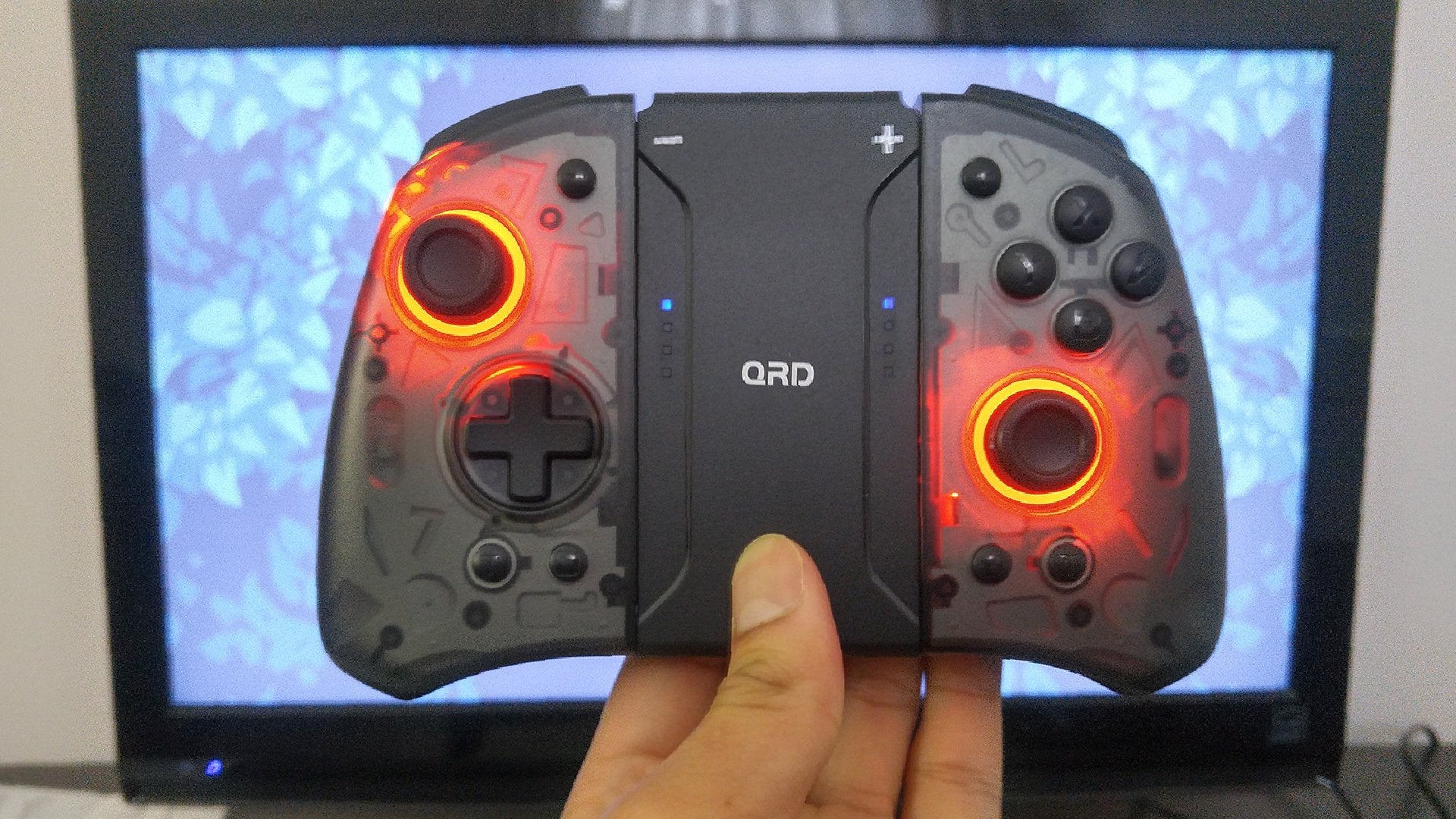 A hand holding up a gray Nintendo Switch controller in front of a monitor. The controller has glowing red lights.